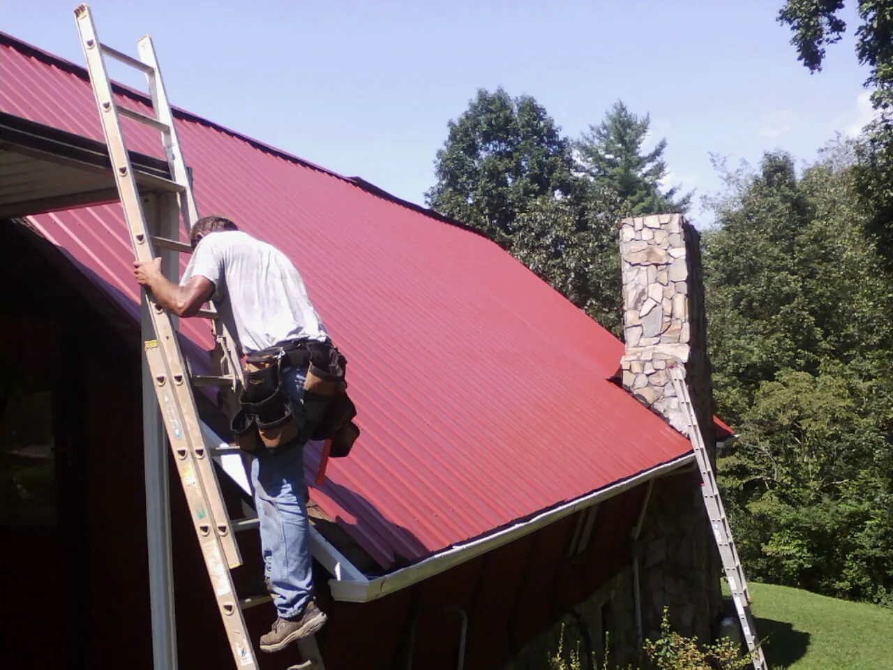 Roofer on a ladder beside a red metal roof and stone chimney — Soffit & Fascia Repair by Roofing Brothers in Willow Street